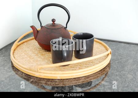 Detail einer roten chinesischen Teekanne und zwei schwarze Tassen auf einem Korbtablett in einer hellen Küche. Niemand drinnen Stockfoto