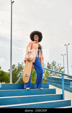 Eine junge Afroamerikanerin mit lockigem Haar steht selbstbewusst auf Stufen und hält ein Skateboard in einem urbanen Skatepark. Stockfoto