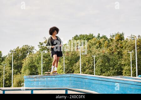 Eine Afroamerikanerin mit lockigen Haaren steht selbstbewusst auf einer Skateboard-Rampe in einem Skatepark im Freien. Stockfoto