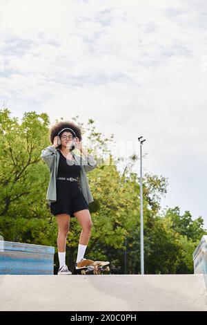 Eine junge Afroamerikanerin mit lockigen Haaren steht selbstbewusst auf einer Skateboard-Rampe in einem Skatepark. Stockfoto