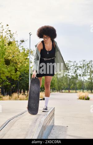 Eine junge afroamerikanische Frau mit lockigen Haaren strahlt Stil aus, während sie in einem Skatepark anmutig ein Skateboard in einem schwarzen Kleid hält. Stockfoto