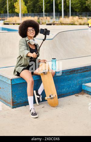 Eine junge Afroamerikanerin mit lockigem Haar, die auf einer Bank mit Skateboard in einem lebhaften Skatepark sitzt. Stockfoto