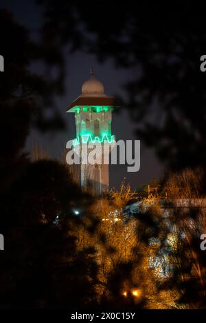 Eine majestätische weiße Moschee mit einem hoch aufragenden, beleuchteten Minarett durchdringt den Nachthimmel, seine Fenster leuchten mit warmem Licht. Nach Dem Ramadan. Grüne Farbe Stockfoto