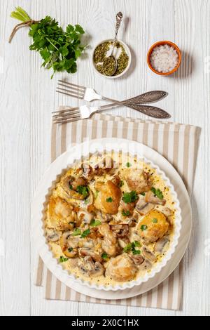 Hähnchenschenkel in einer cremigen Pilz-Knoblauchsauce mit Kräutern und Parmesan in weißer Backform auf weißem Holztisch mit Gabeln, vertikale Ansicht Stockfoto