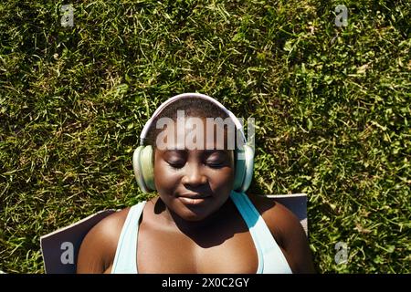 Eine Afroamerikanerin in Sportbekleidung entspannt sich in der Natur und hört Musik über Kopfhörer, während sie im Gras liegt. Stockfoto