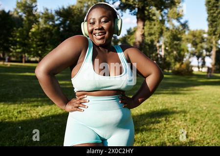 Eine Afroamerikanerin in Sportbekleidung steht im Gras und zeigt sich positiv auf den Körper, während sie Musik über Kopfhörer hört. Stockfoto