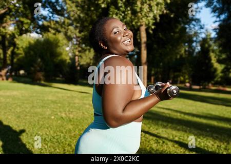 Eine körperpositive Afroamerikanerin in Sportbekleidung hält zwei Kurzhanteln, während sie in einer ruhigen Parklandschaft trainiert. Stockfoto