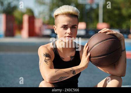 Eine junge Frau mit kurzen Haaren und Tätowierungen, die auf dem Boden sitzt, einen Basketball hält, in Gedanken verloren. Stockfoto