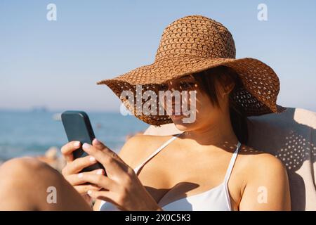 Eine junge Frau mit Strohhut, die an einem sonnigen Tag ein Selfie am Strand macht Stockfoto