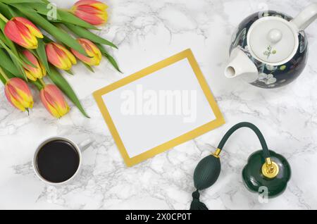 Leeres Blatt Papier mit goldenem Rahmen, umgeben von Tulpen, einer grünen Flasche Parfüm, einer Tasse Kaffee und einer Teekanne. Stockfoto