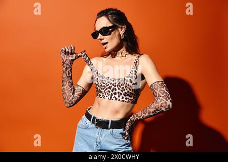 Eine modische Frau trägt selbstbewusst ein Oberteil mit Leopardenmuster und eine Jeans mit Sonnenbrille vor orangefarbenem Studiohintergrund. Stockfoto