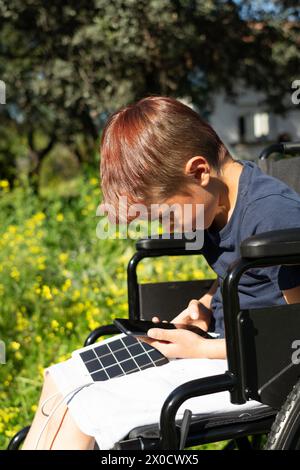 Kaukasischer 8-jähriger Junge mit körperlicher Behinderung im Rollstuhl mit Handy beim Laden mit einem Solarladegerät in der Natur Stockfoto