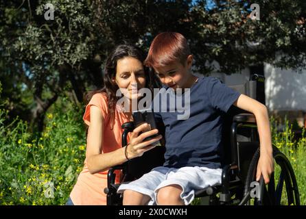 Mutter bringt ihrem Sohn im Rollstuhl etwas auf dem Handy in der Natur bei Stockfoto