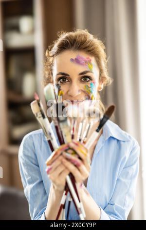 Porträt einer niedlichen Lateinfrau, die einen Haufen Pinsel mit beiden Händen hält und lächelt. Stockfoto