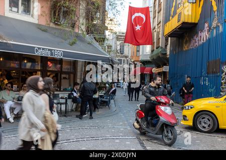 Istanbul, Türkei - 10. April 2024, Menschen sitzen während des Ramadan auf der Veranda eines Cafés im Bezirk Balata. Ein Mann fährt ein Motorrad. Stockfoto