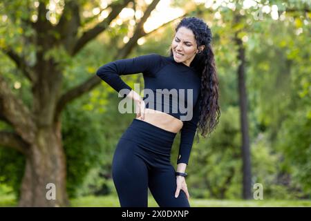 Ein Bild einer hispanischen Frau, die ihren unteren Rücken schmerzhaft hält, während sie im Park steht und sich während eines Workouts unwohl fühlt. Stockfoto