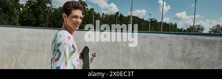 Ein jugendlicher Mann in einem lebhaften Hemd hält an einem sonnigen Tag fröhlich ein Skateboard in einem belebten Skatepark. Stockfoto