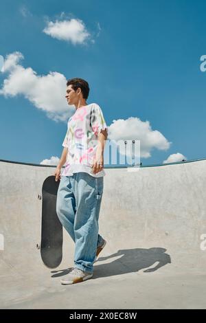 Ein junger Mann hält an einem sonnigen Tag selbstbewusst ein Skateboard in einem pulsierenden Skatepark. Stockfoto