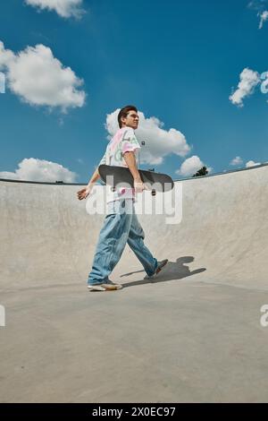 Ein junger Mann läuft lässig mit einem Skateboard in der Hand und strahlt in einem Sommer-Skatepark eine coole und unbeschwerte Atmosphäre aus. Stockfoto