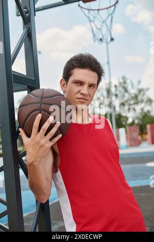 Ein junger Mann in einem roten Hemd hält gekonnt einen Basketball, bereit, an einem Sommertag draußen zu spielen. Stockfoto