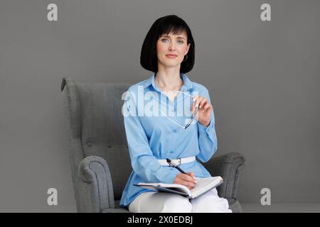 Positive professionelle kaukasische Psychologin schreibt in Papiernotizbuch, schaut und lächelt in die Kamera, arbeitet im Büro mit grauer Wand. Psychothera Stockfoto