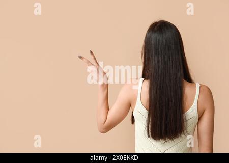 Junge Frau mit Friedenszeichen auf beigefarbenem Hintergrund, Rückansicht Stockfoto