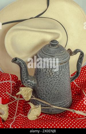 Alte Granitkaffeekanne, ausgefallener Auslauf und Griff, sitzt auf rotem Tuch mit Strohhut Hintergrund. Milchkrautkapseln zeigen ein zwangloses Picknick im Freien an. Stockfoto