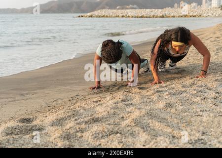 Zwei Frauen absolvieren ein Push-up-Workout an einem Strand mit einer Stadtlandschaft und Bergen in der Ferne, während die Sonne untergeht. Stockfoto