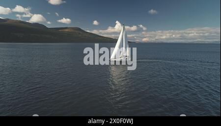 Sonnenspiegelung auf dem Segelboot in der Meereslandschaft. Rennyacht segelt auf offener See. Luxusschiff bei einer Sommer-Bootstour auf Arran Island, Schottland, Europa. Filmisches sanftes Sonnenlicht über dem ruhigen Wasser der Meeresbucht Stockfoto