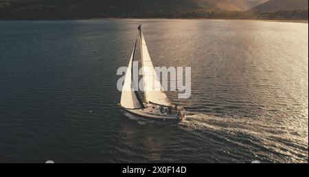 Yachtrundfahrt in Sonnenlicht-Reflexion an der offenen Meeresbucht. Luxussegelboot Segeln am Brodick Pier, Arran Island, Schottland. Mountain isle am Meer. Filmische, ruhige Meereslandschaft mit einsamem Schiff Stockfoto