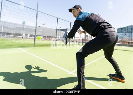 Ein Padel-Spieler springt zum Ball, gut auf der Suche nach Posten und Poster. Mann mit schwarzem Schläger, der draußen hinter dem Netzplatz ein Spiel spielt Stockfoto