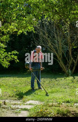 Ein älterer Mann, der mit einer Freischneide Gras in seinem ländlichen Garten in zala, ungarn, abschneidet Stockfoto