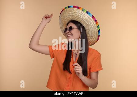 Junge Frau mit mexikanischem Sombrero-Hut und Sonnenbrille, die auf beigefarbenem Hintergrund tanzt Stockfoto