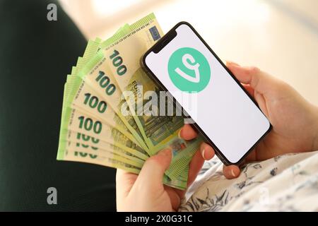 KIEW, UKRAINE - 1. APRIL 2024 Weinstock-Symbol auf dem Smartphone-Bildschirm und Geld in weiblicher Hand. IPhone-Display mit App-Logo und hundert Euro-Scheine in Mädchen Hand nah dran Stockfoto