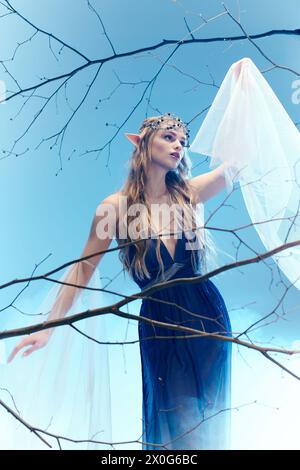 Eine junge Frau in einem fließenden blauen Kleid, die einer Elfenprinzessin ähnelt, steht anmutig auf einem Baum auf eine skurrile und märchenhafte Weise. Stockfoto