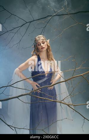 Eine junge Frau in einem blauen Kleid steht anmutig vor einem majestätischen Baum und verkörpert eine märchenhafte Präsenz in einem Studio-Setting. Stockfoto