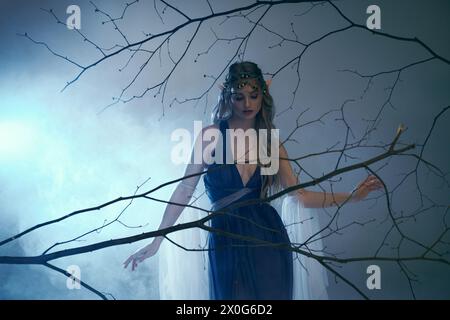 Eine junge Frau mit Elfenprinzessinnen-Vibes steht anmutig vor einem Baum und trägt ein atemberaubendes blaues Kleid. Stockfoto