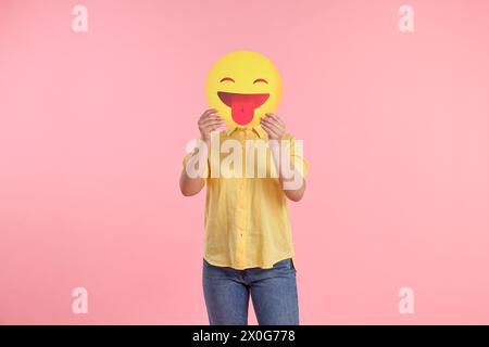 Frau, die das Gesicht mit Emoticon bedeckt und die Zunge auf rosa Hintergrund hinausragt Stockfoto