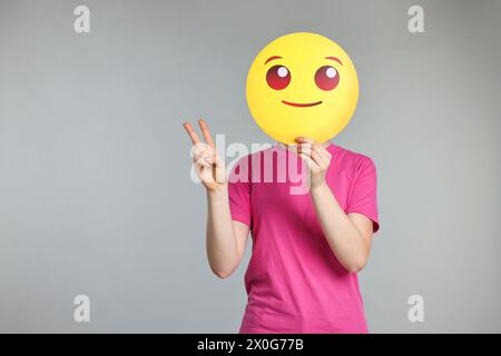 Frau, die das Gesicht mit lächelndem Emoticon bedeckt und Friedenszeichen auf grauem Hintergrund zeigt. Leerzeichen für Text Stockfoto