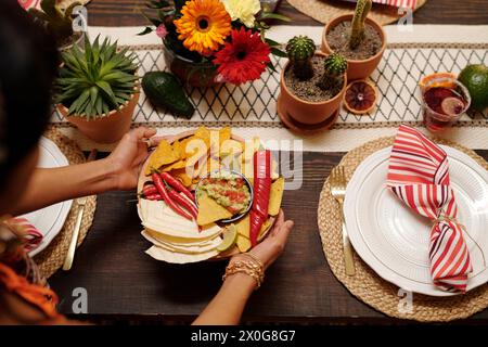 Hände einer jungen hispanischen Frau, die appetitliche Chips, Quesadillas, rote heiße Chili-Paprika und scharfe hausgemachte Soße auf festlichen Tisch legt Stockfoto