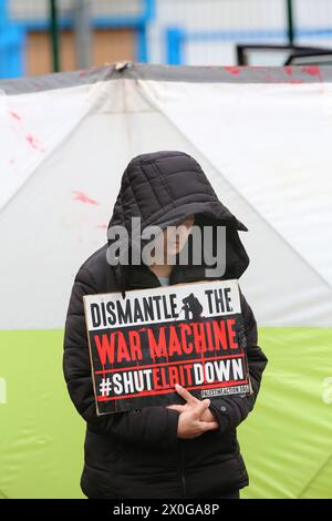 Ein Demonstrant steht mit einem Schild mit der Aufschrift "Zerlegen der Kriegsmaschine - Schließen Sie Elbit herunter", während Polizisten daran arbeiten, hinter einem tragbaren Bildschirm vor der Fabrik von Elbitís Aktivist freizulassen. Zwei Aktivisten der Palästinensischen Aktion fahren zwei Autos in die Poller vor den Toren der Fabrik von Elbitís und verriegelten sich mit einem D-Lock um den Hals an den Lenkrädern, um zu verhindern, dass Arbeiter den Standort betreten. Die Fabrik für das unbemannte Luftfahrzeug-Taktische System (UTACS) gehört dem israelischen Rüstungsunternehmen Elbit Systems. Es ist eines von sieben Einrichtungen, die Elbit im Vereinigten Königreich Waffen und Drohnen für die Israe herstellt Stockfoto