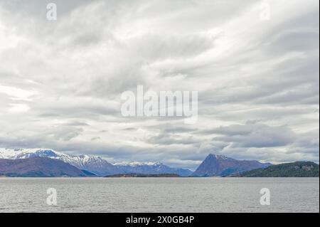Verschneite Gipfel ragen über dem ruhigen Meer, unter einem bewölkten Himmel, in einer ruhigen Landschaft. Stockfoto