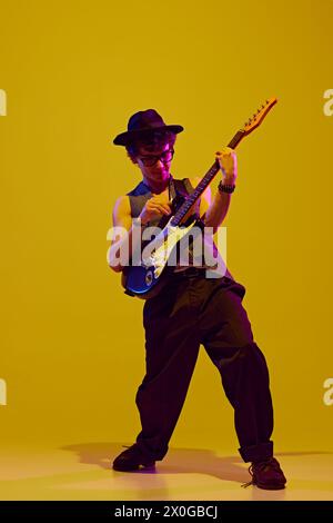 Porträt eines jungen charismatischen, talentierten Mannes, der Rock-n-Roll in Neonlicht vor gelbem Studiohintergrund spielt. Stockfoto