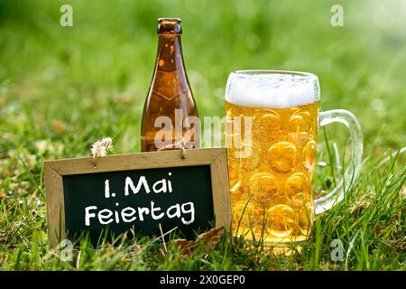 12. April 2024: Plakette auf einer grünen Wiese mit einem Bierkrug und einer Bierflasche mit der Aufschrift: Feiertag 1. Mai. FOTOMONTAGE *** Tafel in einer grünen Wiese mit einem Bierkrug und einer Bierflasche mit Aufschrift: 1. Mai Feiertag. FOTOMONTAGE Stockfoto