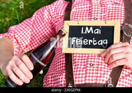 12. April 2024: Mann in traditioneller Tracht auf einer Wiese mit Bier und Brett mit der Inschrift: 1. Mai Feiertag. FOTOMONTAGE *** Mann liegt in Tracht in einer Wiese mit einem Bier und einer Tafel mit der Aufschrift: 1. Mai Feiertag. FOTOMONTAGE Stockfoto