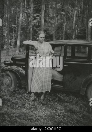 Eine junge Frau, etwa 22 Jahre alt, lehnt sich an die weit geöffnete Fahrertür eines großen Autos (Horch?) Mit erhobenem Arm. Sie trägt ein wadenlanges, leichtes kariertes Kleid mit Ellbogenlangen Ärmeln. Sie hat ihr kurzes blondes Haar zurückgekämmt. Das Auto steht in einem kargen Fichtenwald, wobei der linke Vorderreifen fast den Baum berührt. Der Waldboden ist knöchelhoch mit Blaubeerbüschen bewachsen. Die Frau schaut angespannt, erwartungsvoll von der Kamera weg zu einem Ereignis, das sich hinter ihr versteckt [automatisierte Übersetzung] Stockfoto