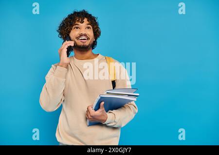 Ein indischer Student in lässiger Kleidung steht vor blauem Hintergrund, hält eine Mappe und spricht mit einem Handy. Stockfoto