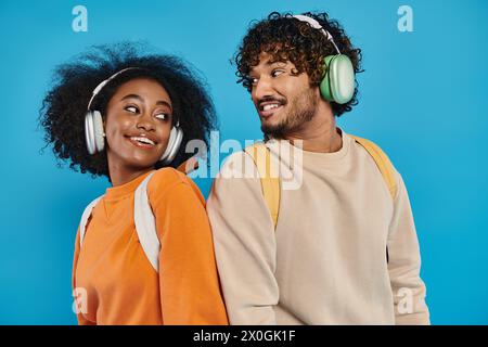 Ein interrassisches Paar steht zusammen, trägt Kopfhörer und teilt Musik vor blauem Hintergrund in einem Studio. Stockfoto