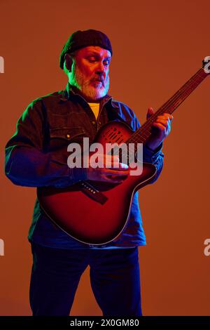 Leidenschaftlicher Senior-Gitarrist aus Denim in farbenfrohem Neonlicht vor orangefarbenem Studiohintergrund. Stockfoto