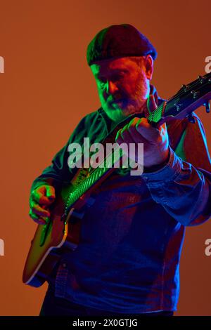 Gitarrist mit Bart passt E-Gitarre in farbenfrohem Neonlicht vor orangefarbenem Studiohintergrund an. Stockfoto